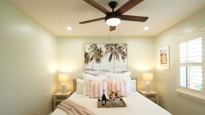 A bedroom with a bed, two bedside lamps, a ceiling fan, a beach-themed wall art, and a window with blinds. - Turfside Terrace, Heated Pool and Putt Putt (Jupiter)