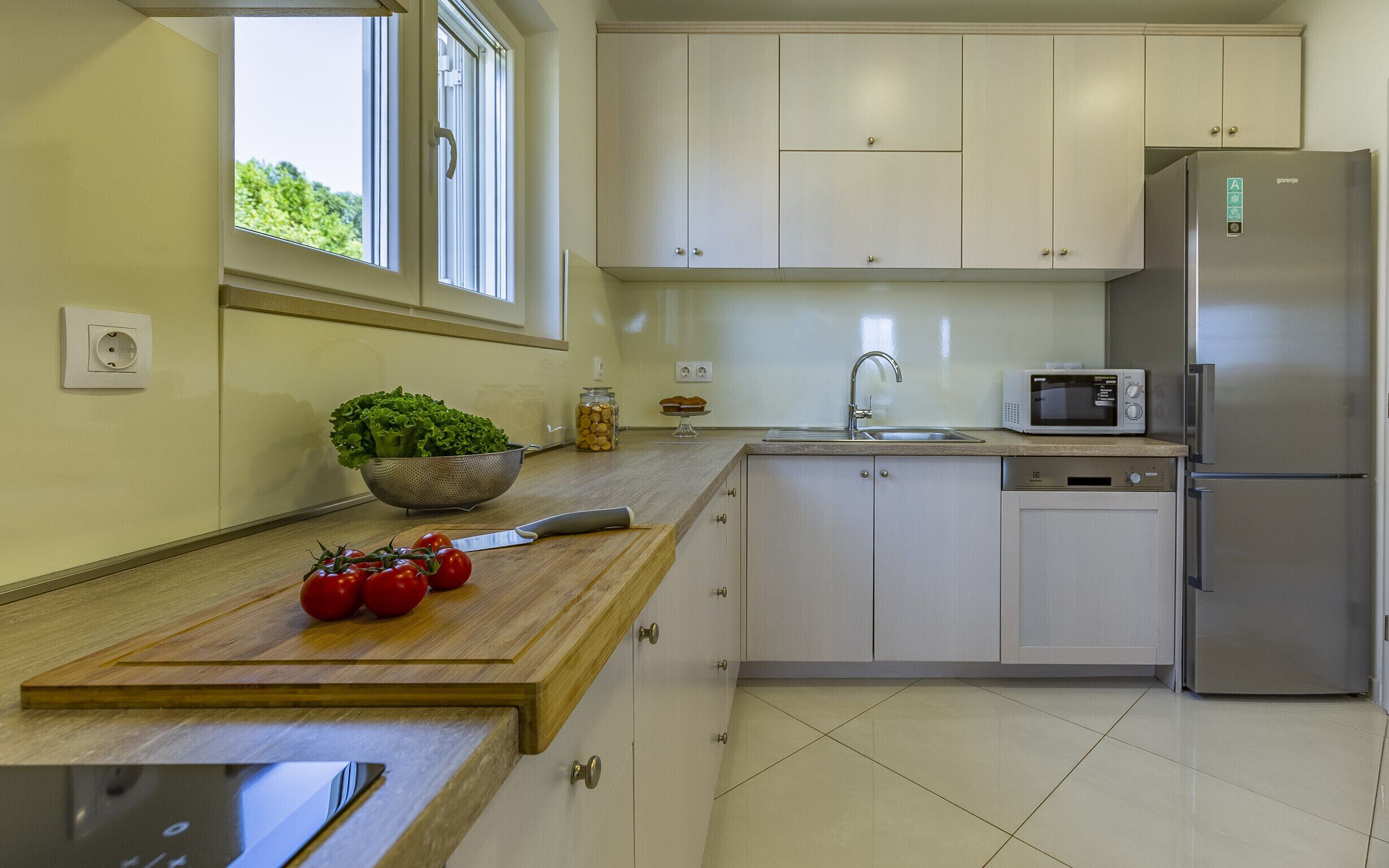 Una cocina moderna con armarios blancos, un refrigerador de acero inoxidable, un microondas y una encimera de madera con tomates frescos y un colador.