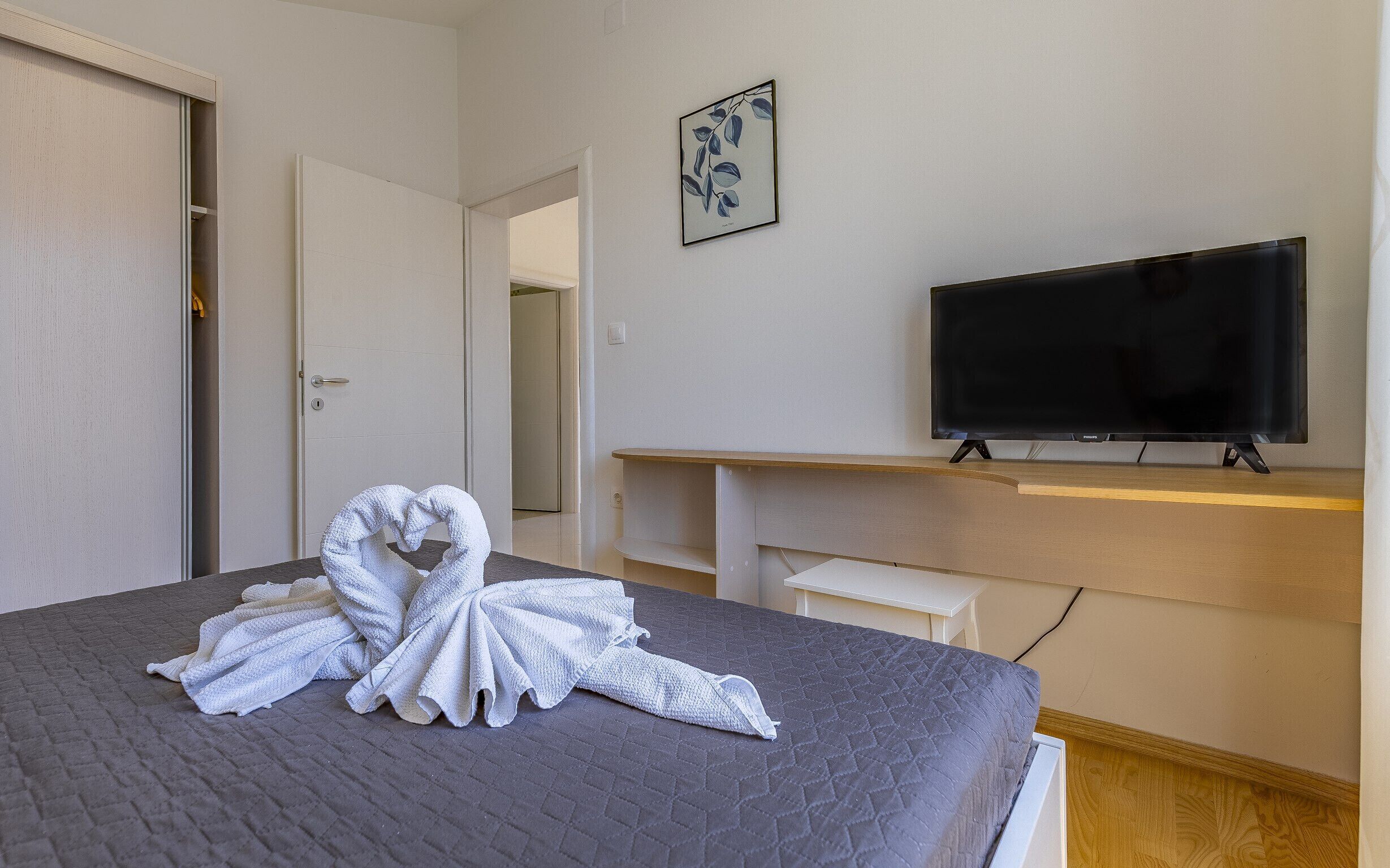 Ein Hotelzimmer mit Bett, Fernseher, einem Bild an der Wand und einem herzförmigen Handtuch auf dem Bett.