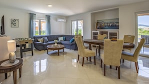 A modern living room with a sofa, coffee table, dining table, and chairs. There is a foosball table, a painting on the wall, and a window with curtains. - Lotus Elite Villa with Pool & Jacuzzi 1 (Pula)