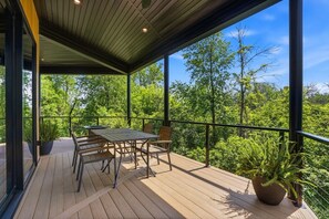 Een overdekt terras buiten met een tafel en stoelen, omgeven door groen en bomen.