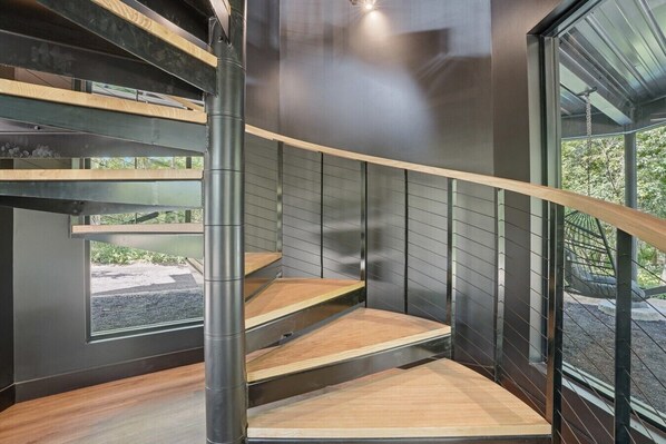 A modern interior with a spiral staircase, metal railings, and a view of greenery outside.