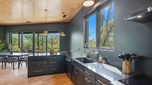 A modern kitchen with dark cabinetry, a black countertop, and a large window providing a view of the outdoors. - The Outlook Treehouse & Lavender Farm (Oxford)