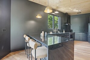 A modern kitchen with a large island, bar stools, pendant lighting, and a view of greenery outside.