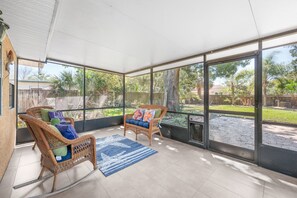 Un patio cubierto con sillas de mimbre y un tapete azul, ventanas amplias y vista a un jardín.