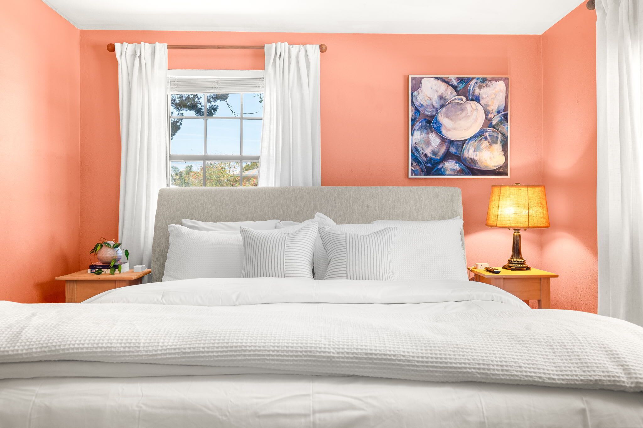 A bedroom with a bed, a nightstand with a lamp, a window with curtains, and a painting on the wall.