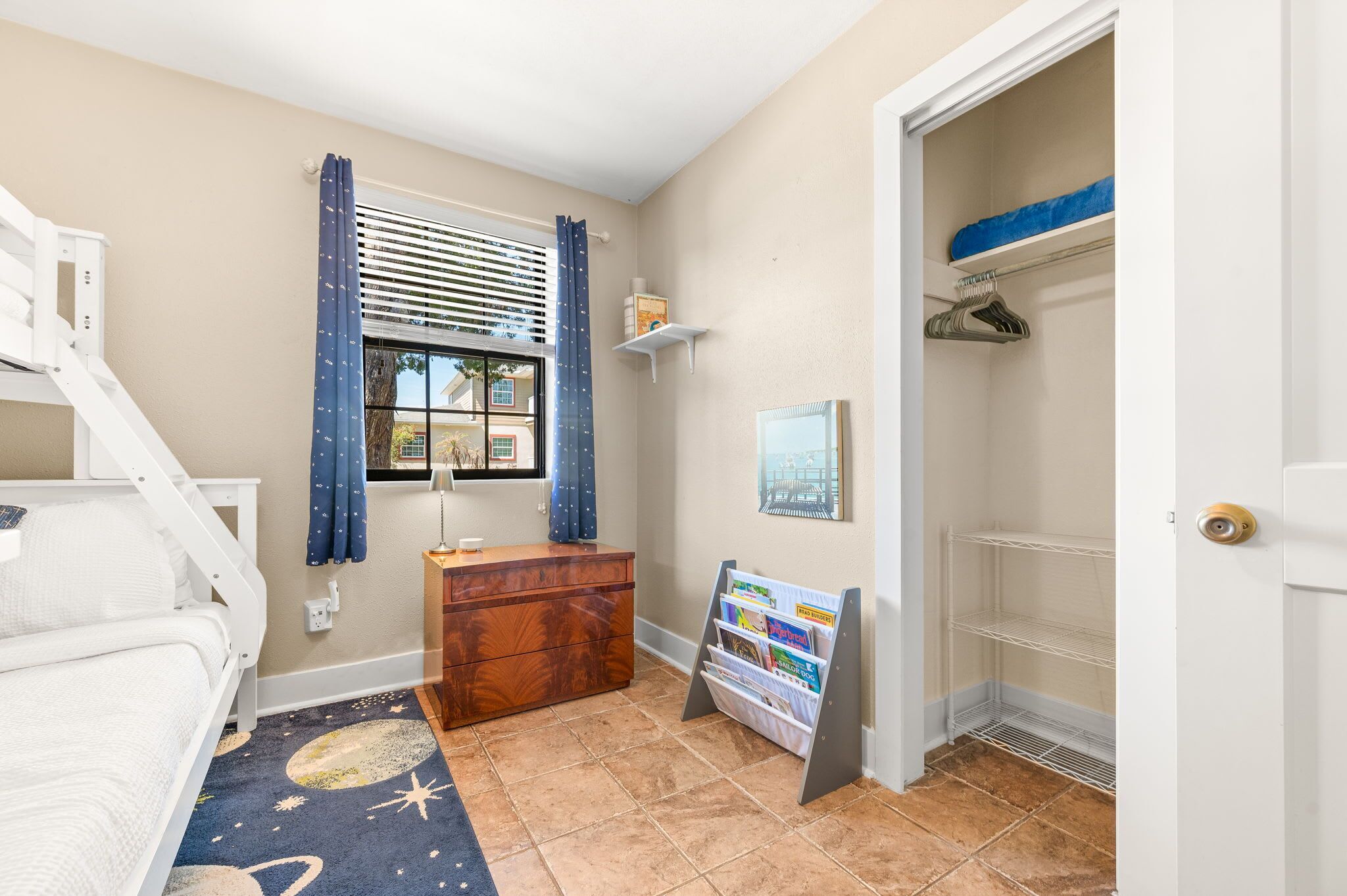 Une chambre à coucher comprenant un lit, une commode, une table de chevet, une fenêtre avec des rideaux, une étagère avec des livres et une porte avec un porte-manteau.