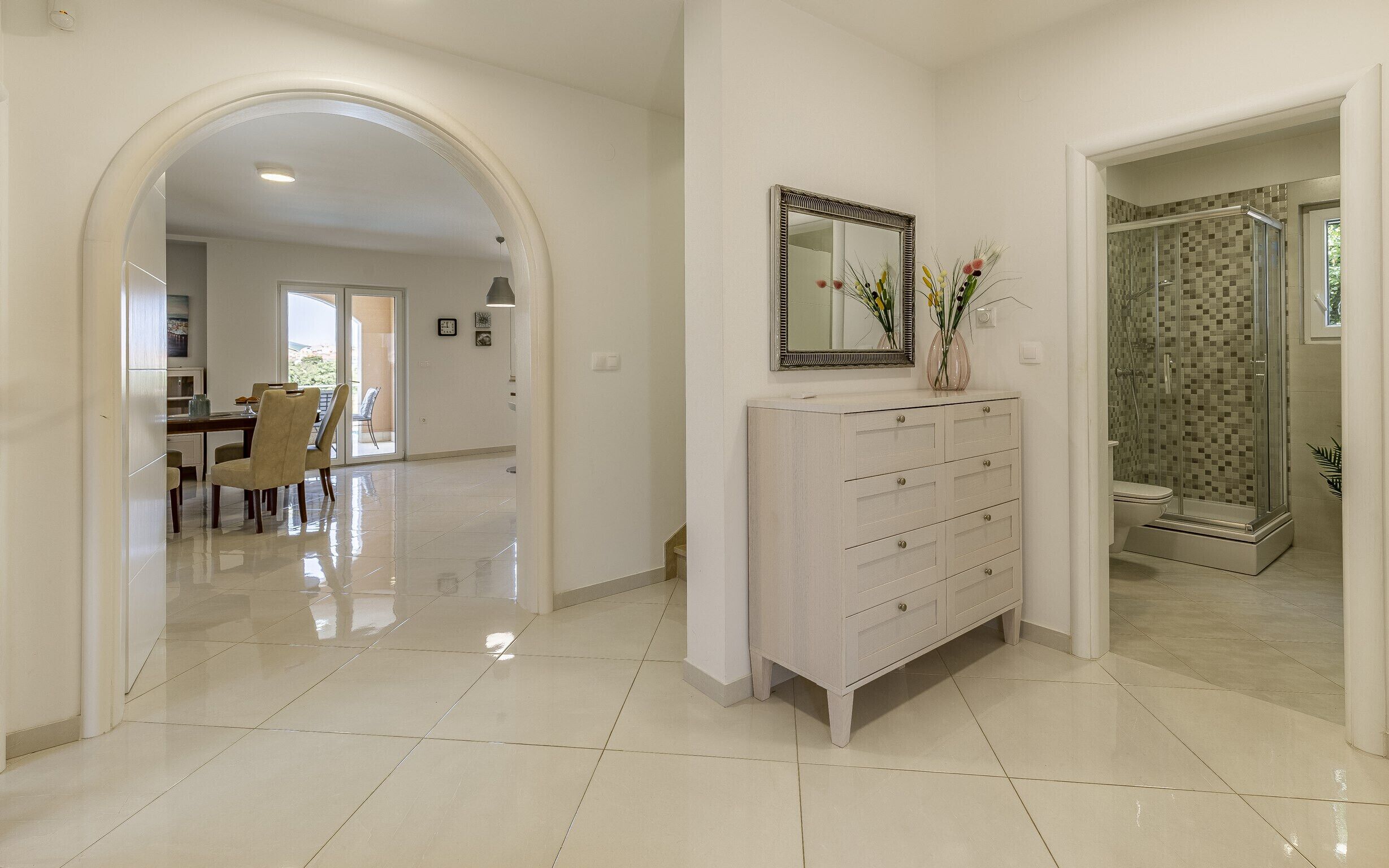 A spacious hallway with a dining area, a white console with drawers, and a shower area with mosaic tiles.