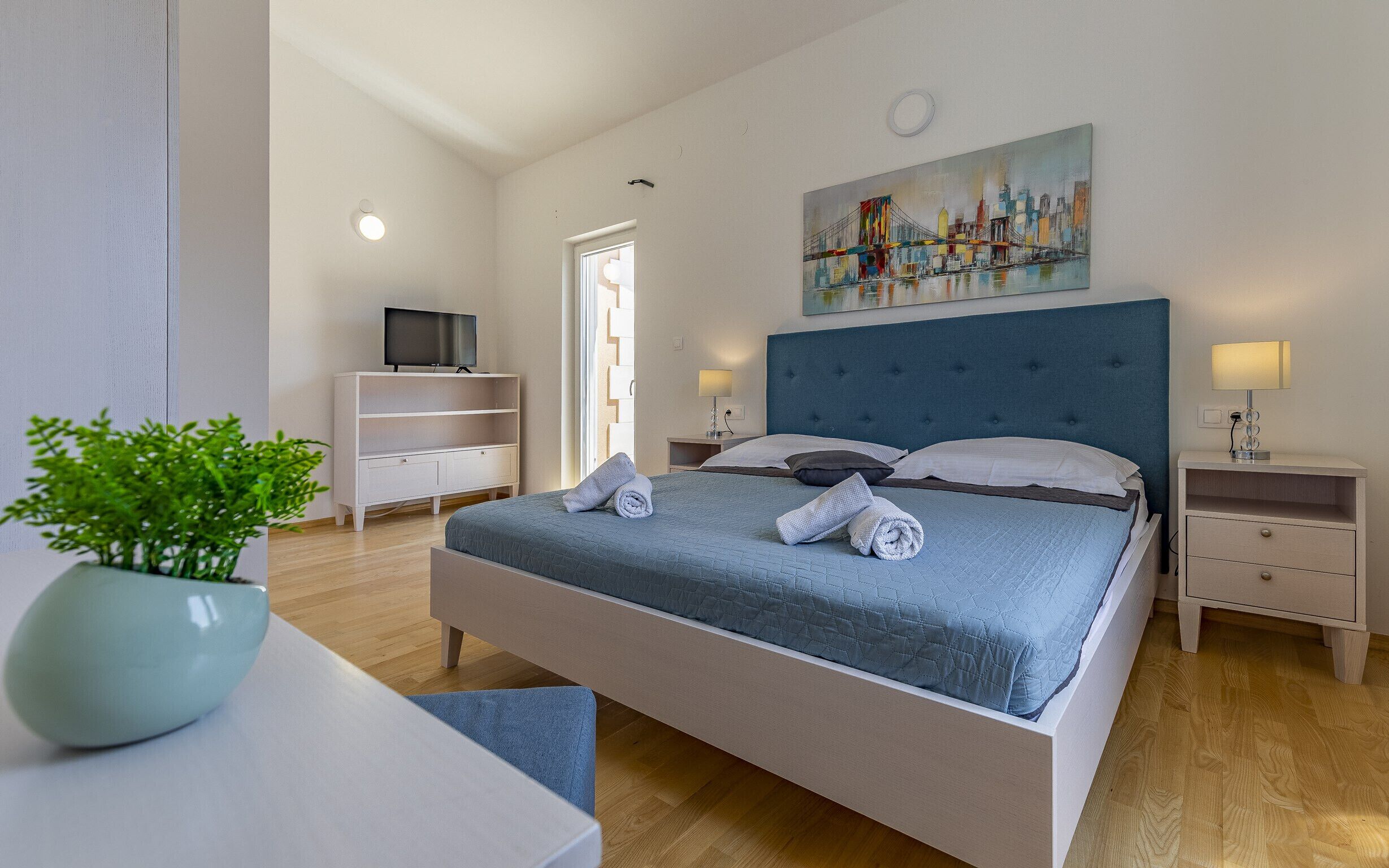 Camera d'hotel moderna con un letto grande, una TV su un mobile bianco, un quadro appeso al muro e una pianta in vaso.