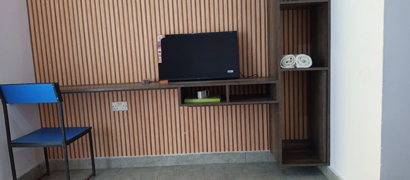 A room with a wooden slatted wall, a television on a shelf, a blue chair, and a small shelf with rolled-up items.