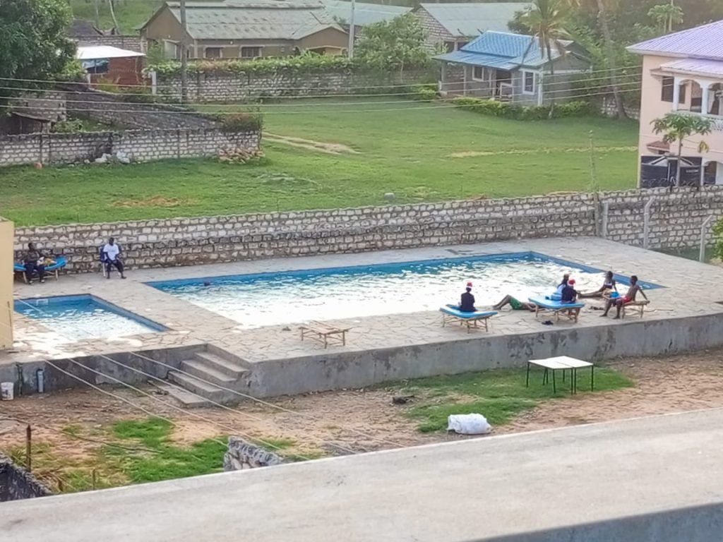 A swimming pool area with a stone wall, a grassy field, and houses in the background.
