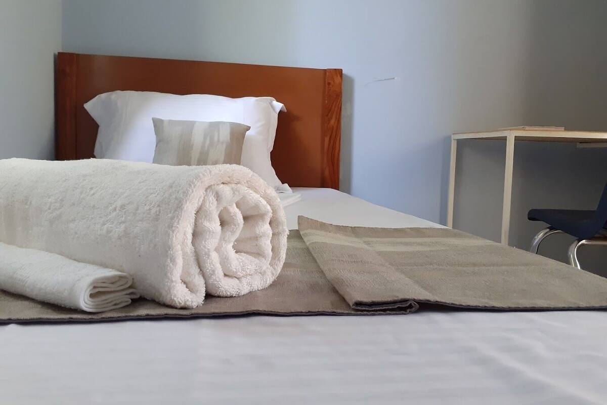 Ein ordentlich bezogenes Bett mit einem aufgerollten weißen Handtuch und einer beigen Decke, ein hölzernes Kopfteil und ein kleiner Schreibtisch mit Stuhl in einem Hotelzimmer.