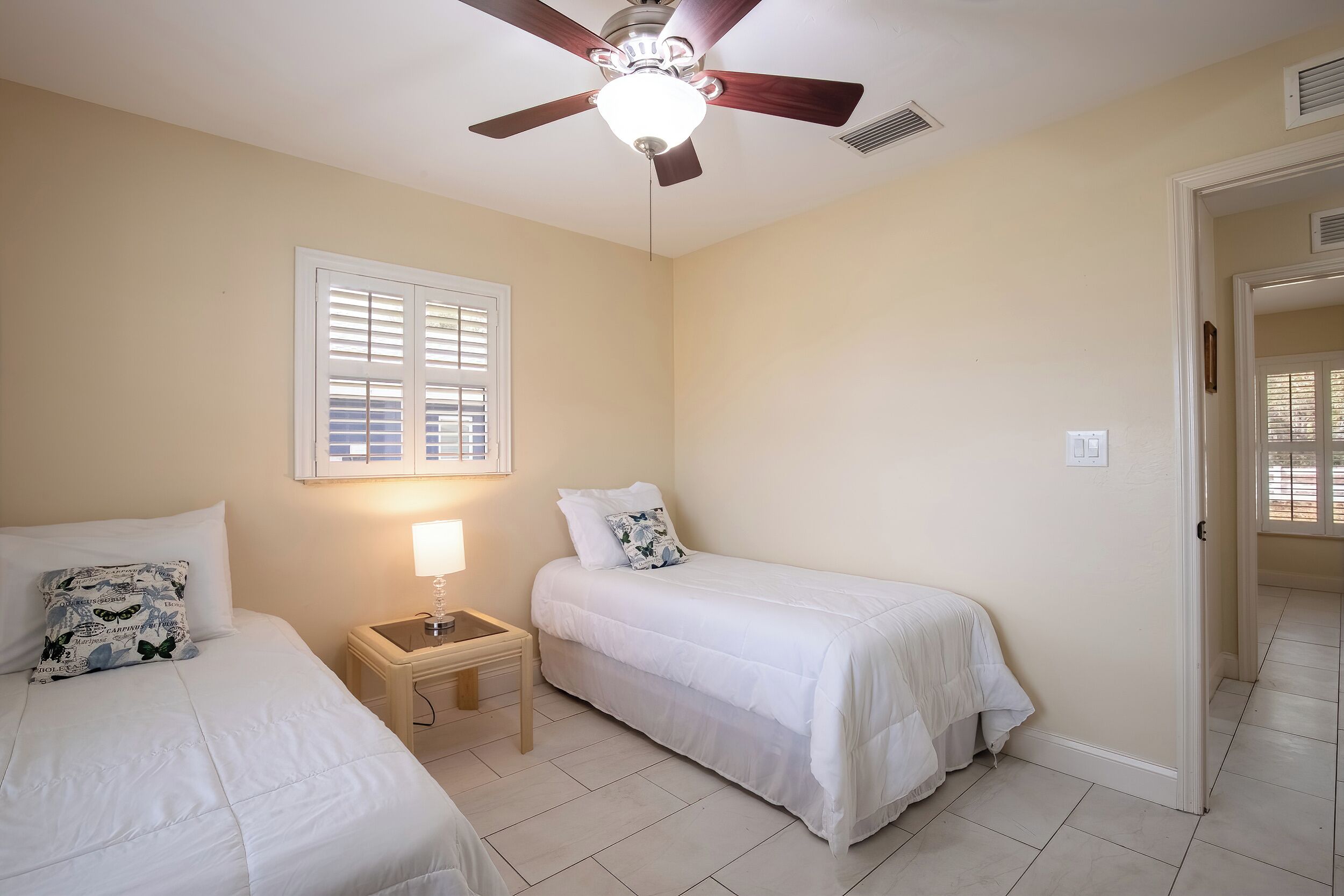 A bedroom with two beds, a ceiling fan, a window with blinds, a bedside table with a lamp, and a door leading to another room.