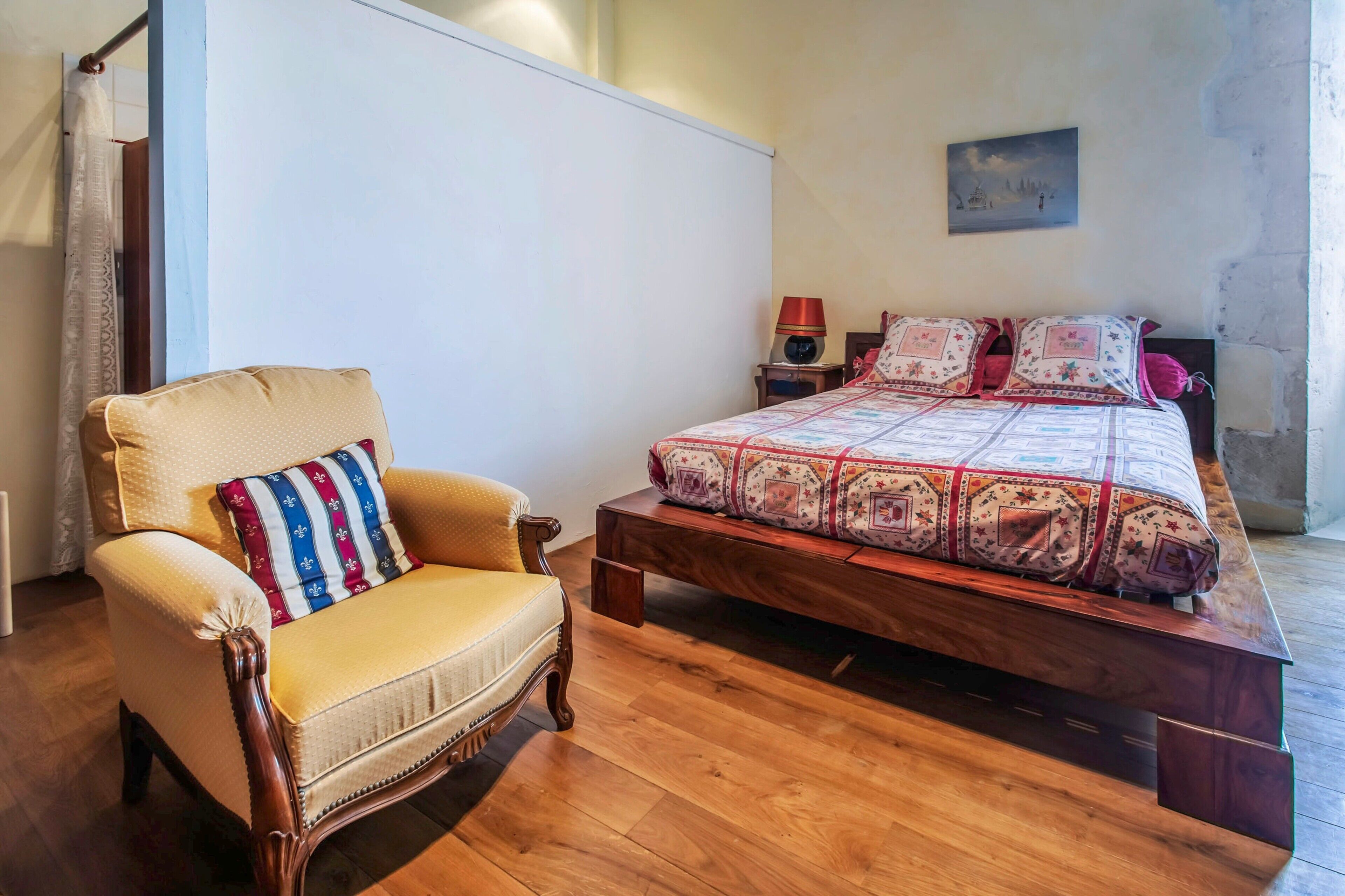 Un dormitorio con una cama de madera, un sillón tapizado, una mesita de noche y una lámpara de pared.