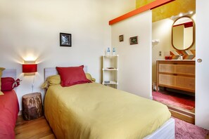 Quarto com cama, mesas de cabeceira, um aparador, guarda-roupa, espelho, prateleira com garrafas e luminária na parede.