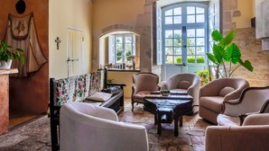 Ein gemütlicher Raum mit Steinfußboden, einem großen Bogenfenster und verschiedenen Sitzgelegenheiten, darunter eine Couch, Stühle und ein kleiner Tisch.
