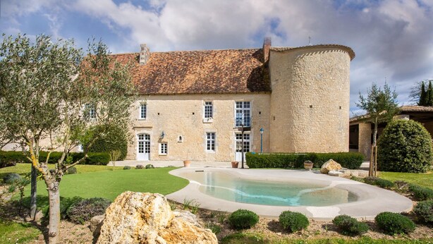 En byggnad i sten med en swimmingpool, omgiven av en välskött trädgård och en stor sten.