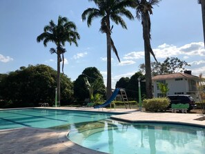 A swimming pool with a blue slide, surrounded by palm trees and a white building.