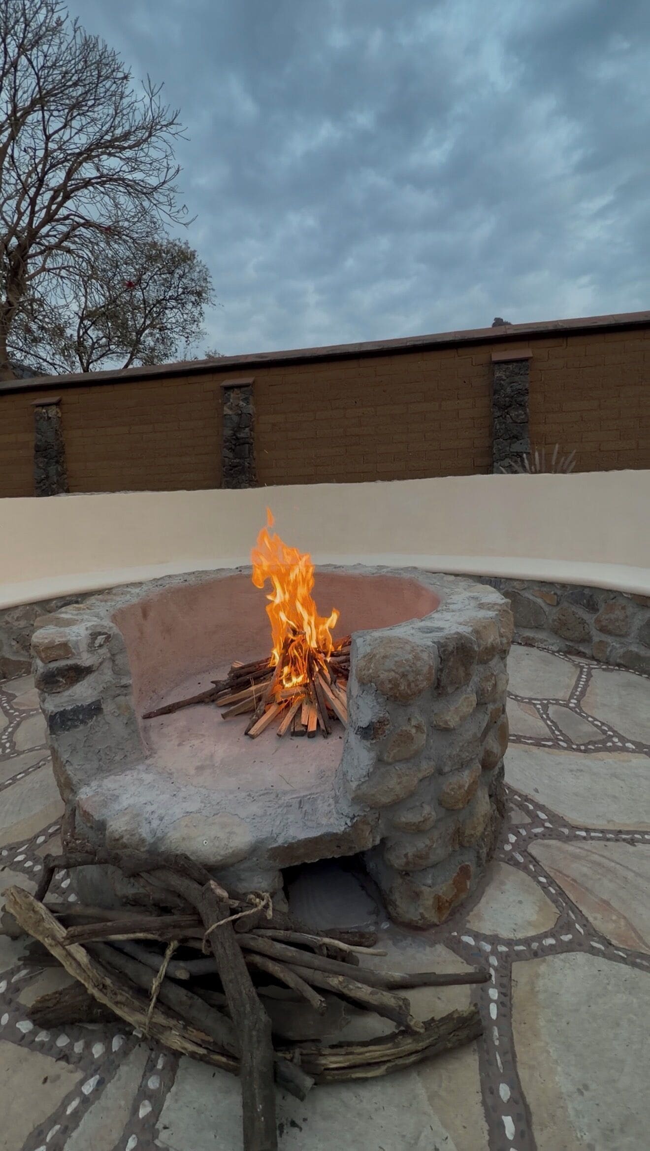 A stone fire pit with a fire burning in the center, surrounded by a paved patio with a decorative pattern.
