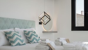 Ein ordentlich bezogenes Bett mit weißen Leinen und einem Kissen in Türkis und Weiß mit Zickzackmuster, eine wandmontierte geometrische Leuchte und ein Stapel gefalteter Handtücher.