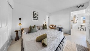 A modern bedroom with a large bed, a small side table, a chair, and a view into the kitchen.