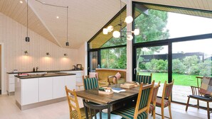A modern kitchen with a central island, dining area with wooden chairs and a table, and large windows offering a view of the outdoors. - 4 Star Holiday Home in Gilleleje (Gilleleje)