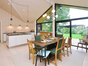 A modern kitchen with a central island, dining area with wooden chairs and a table, and large windows offering a view of the outdoors. - 4 Star Holiday Home in Gilleleje (Gilleleje)