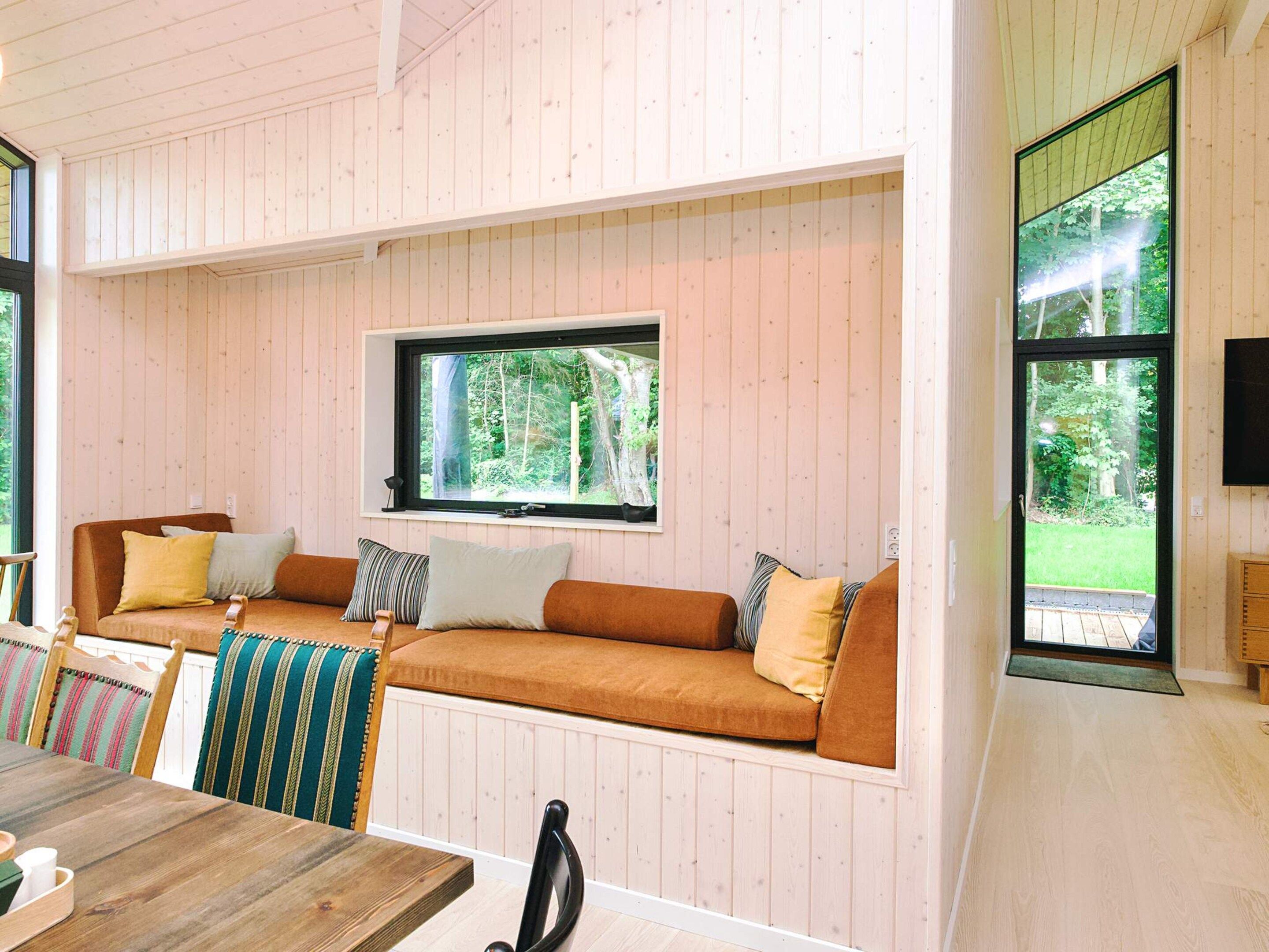 A cozy cabin interior with a wooden interior, a large window with a forest view, a sofa with cushions, and a dining table with chairs.