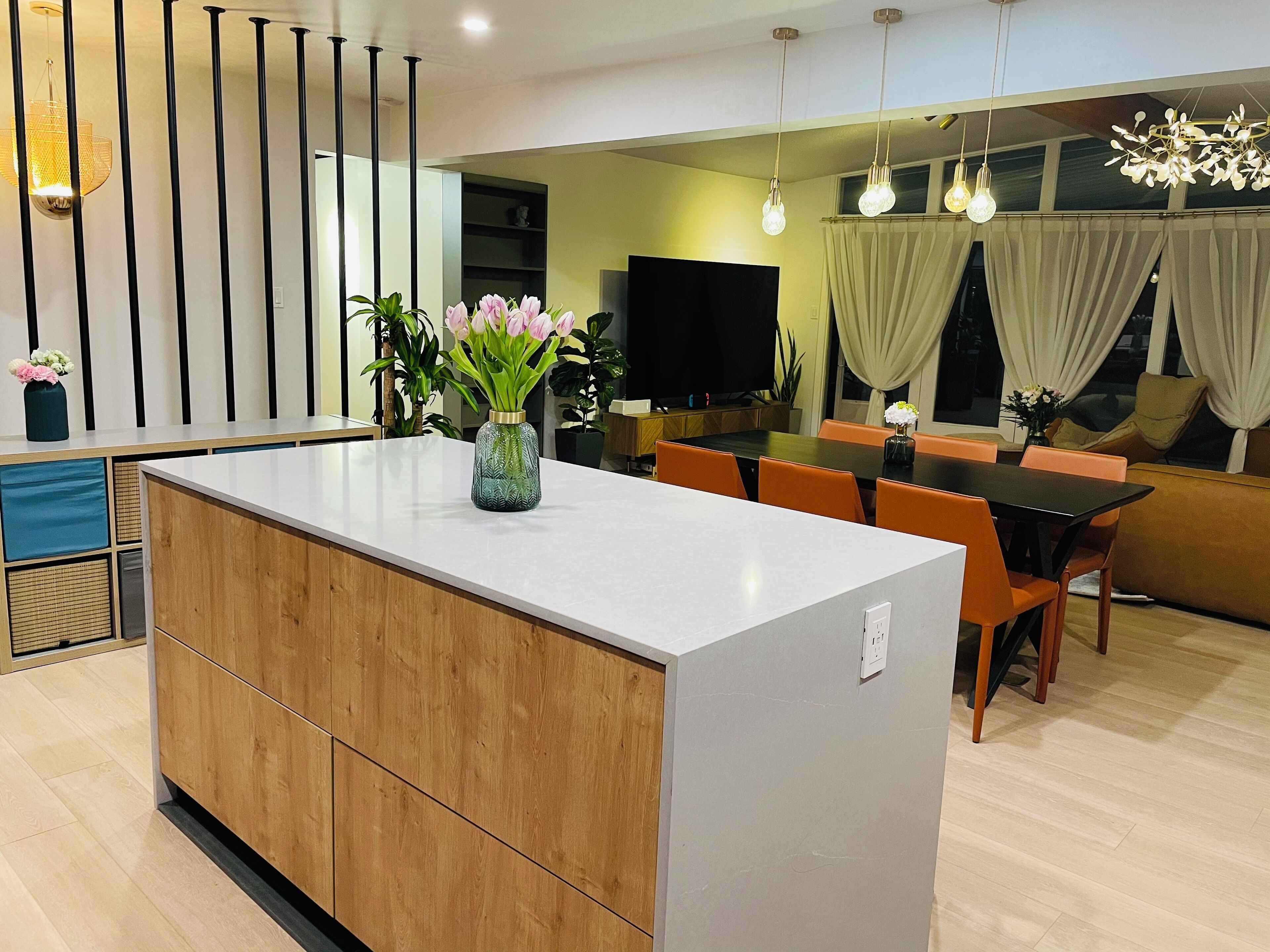 Eine moderne Küche mit einer zentralen Kochinsel, Holzschränken und einem Essbereich mit orangefarbenen Stühlen und einem schwarzen Tisch.