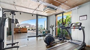 A gym with treadmills, elliptical machines, and weight equipment. It has large windows with a view of a cityscape and a mountain.