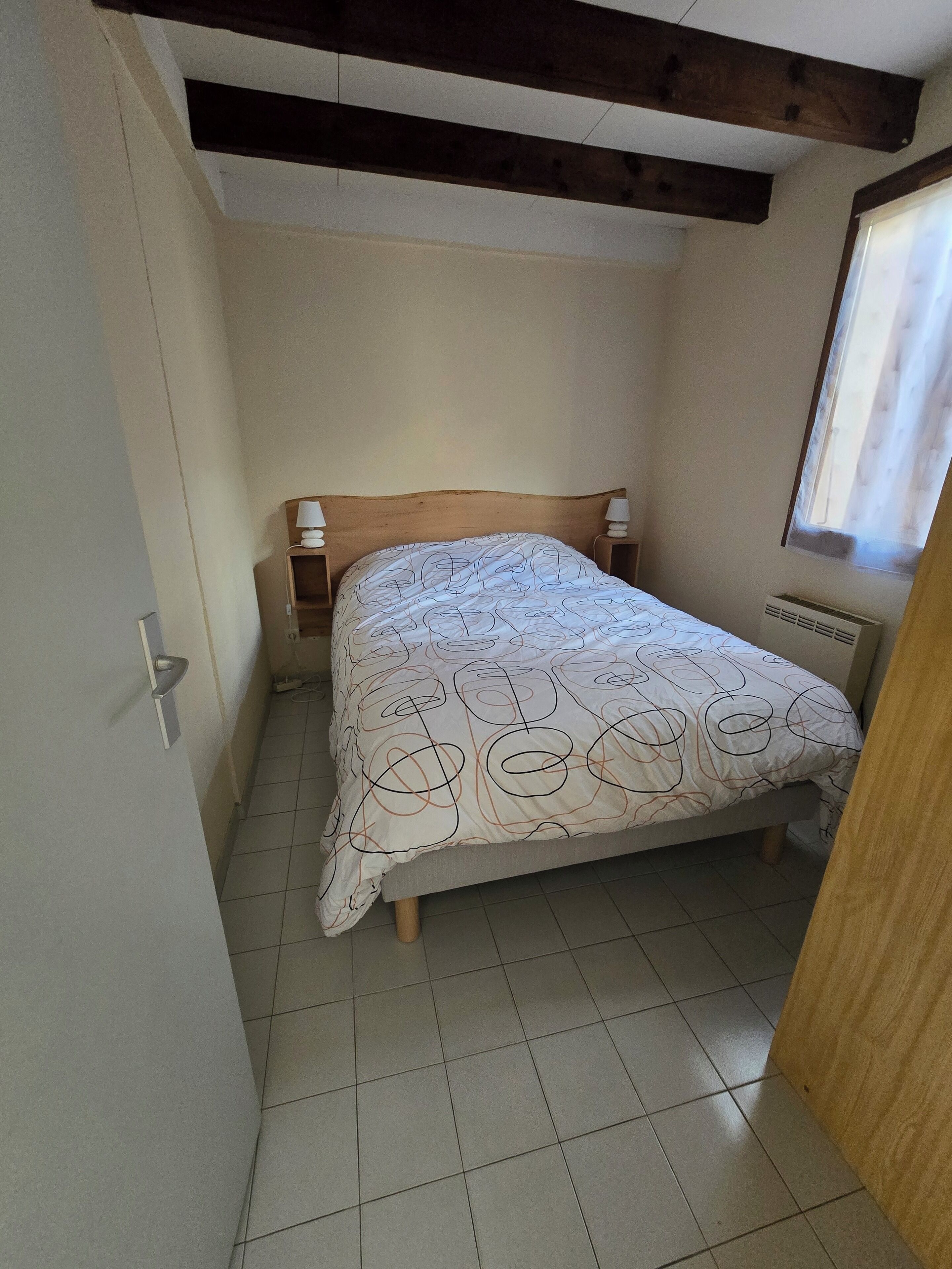 A small, narrow room with a single bed, a wooden headboard, and a window with a curtain.
