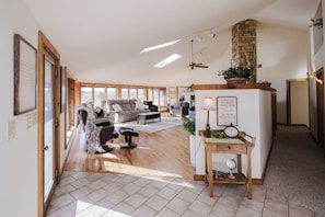 A spacious living room with a large sofa, a coffee table, and a fireplace. There is a wooden table with a clock and a sign on it. The room has large windows and a high ceiling with skylights.