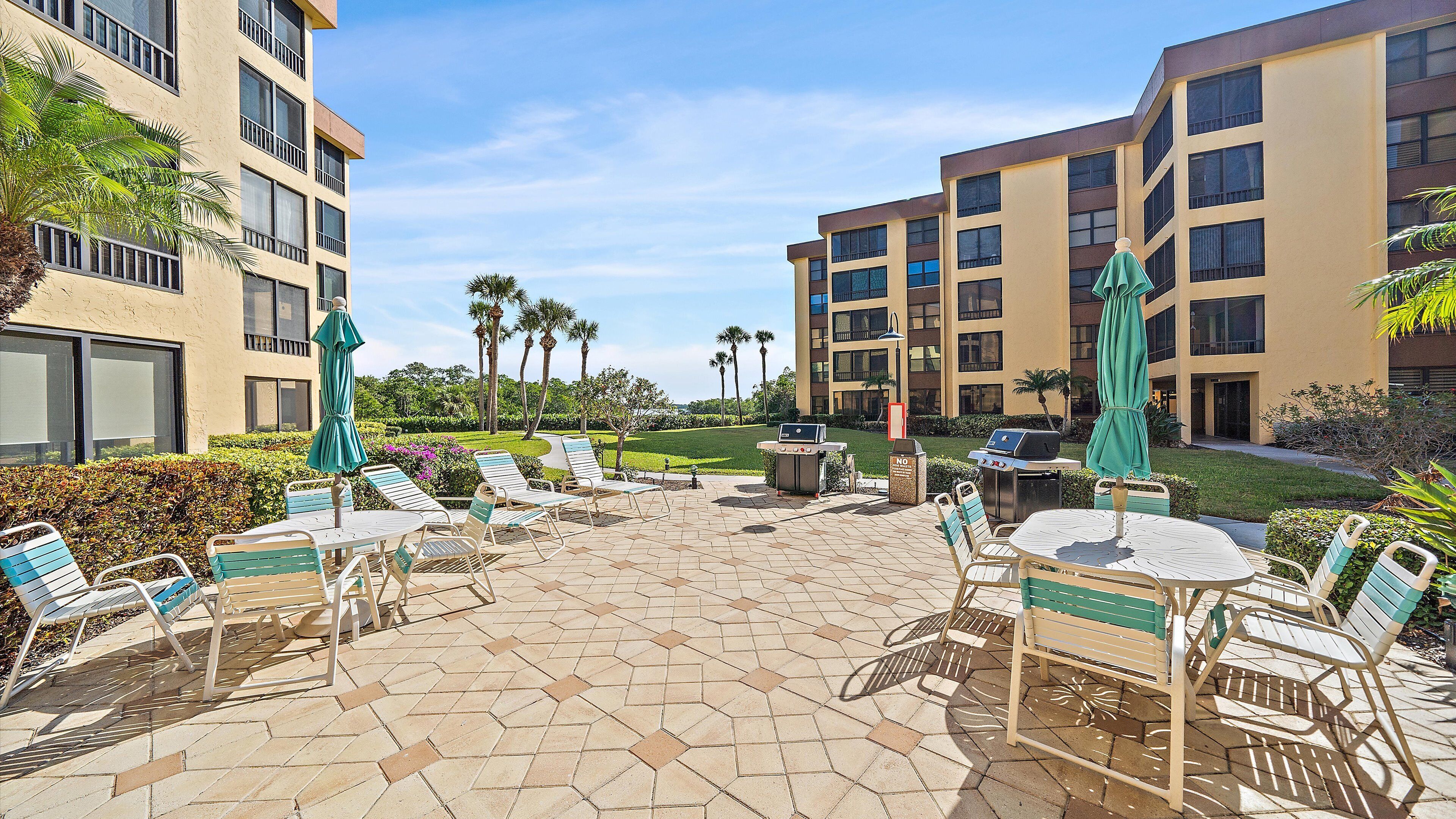 An outdoor patio area with lounge chairs and tables, a grill, and a view of a landscaped garden with palm trees.