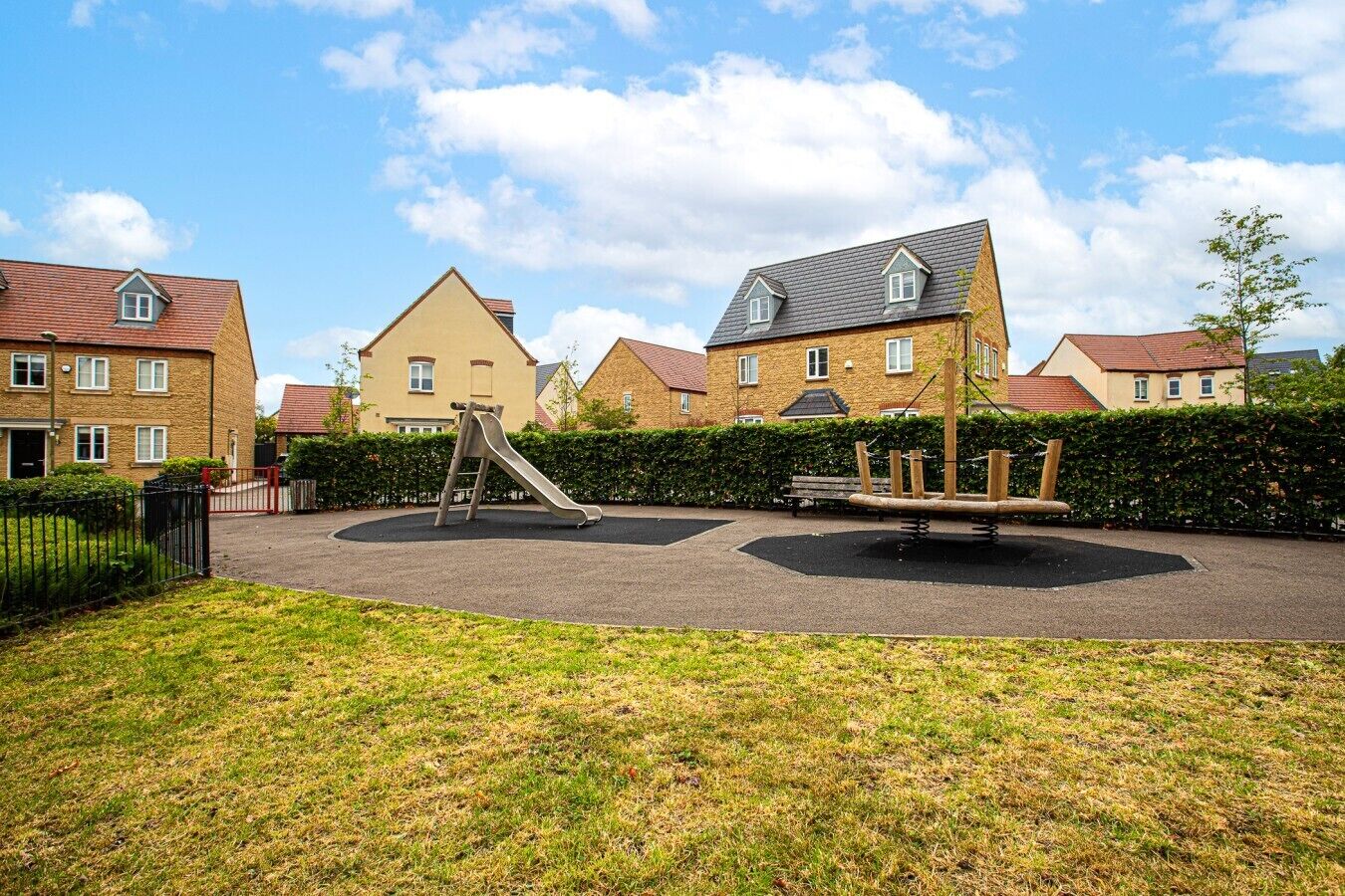 A playground with a slide and swings, surrounded by a hedge and residential buildings.