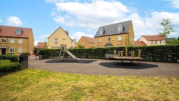 A playground with a slide and swings, surrounded by a hedge and residential buildings.