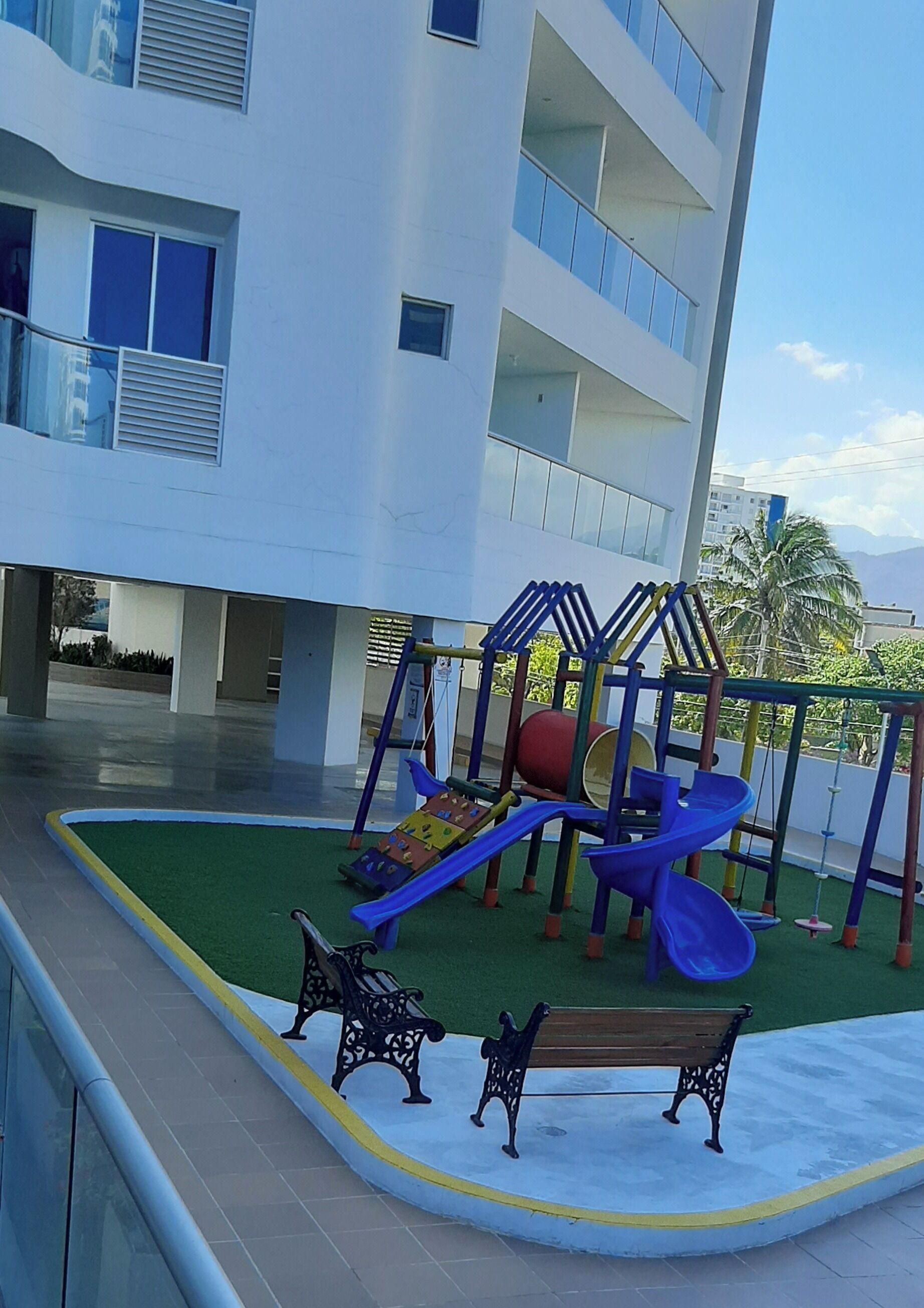 A playground with slides and climbing structures, benches, and a building with balconies in the background.