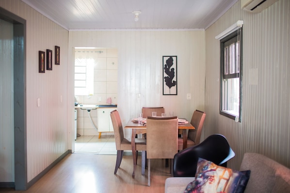 A dining area with a table and chairs, a kitchen in the background, and a window with blinds.