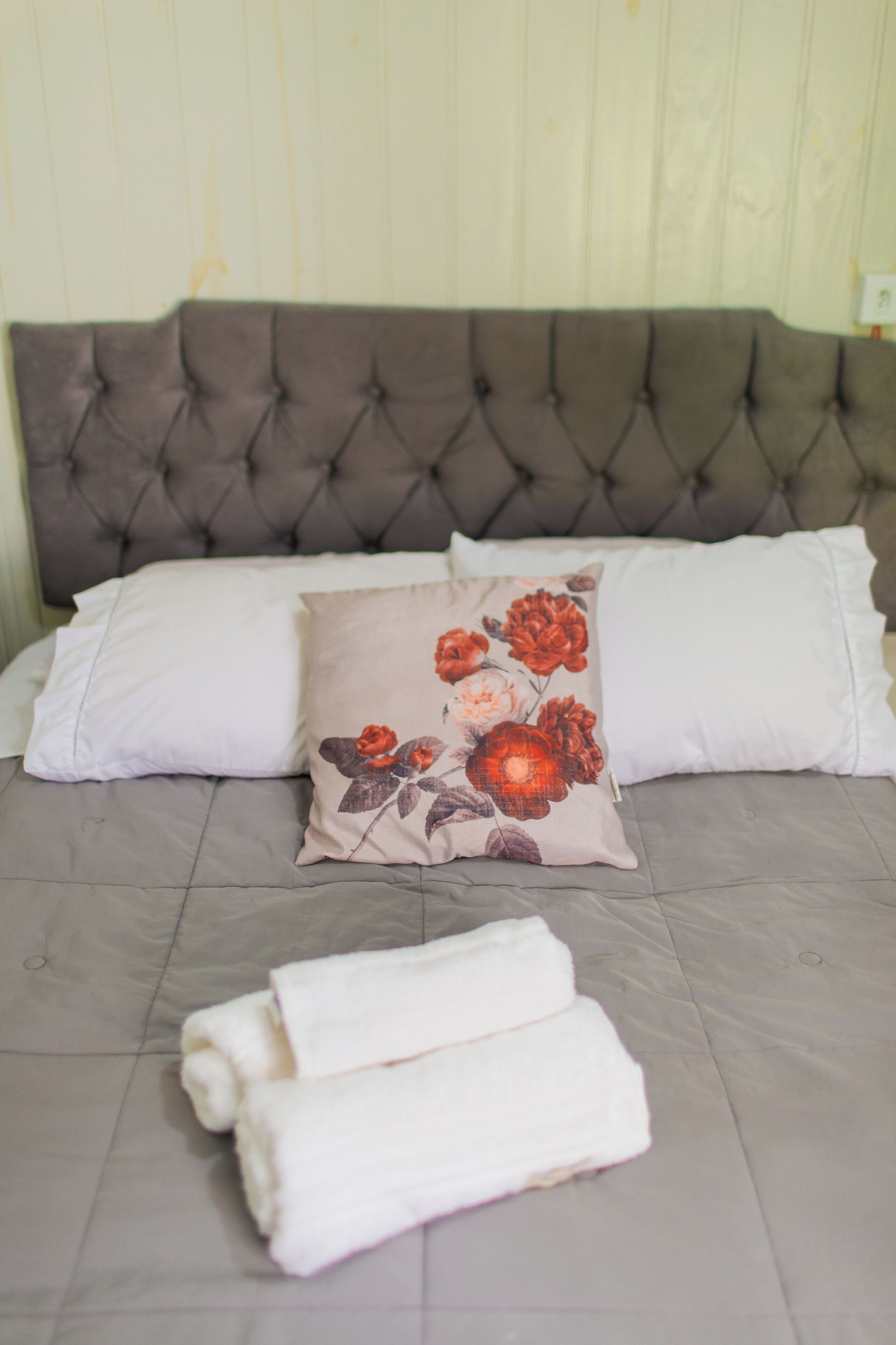 Una cama con cabecera capitonada, dos almohadas blancas, una almohada estampada con flores y dos toallas enrolladas.