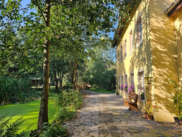 Un chemin de pierres mène à un bâtiment à la façade jaune, entouré de verdure et d’arbres.