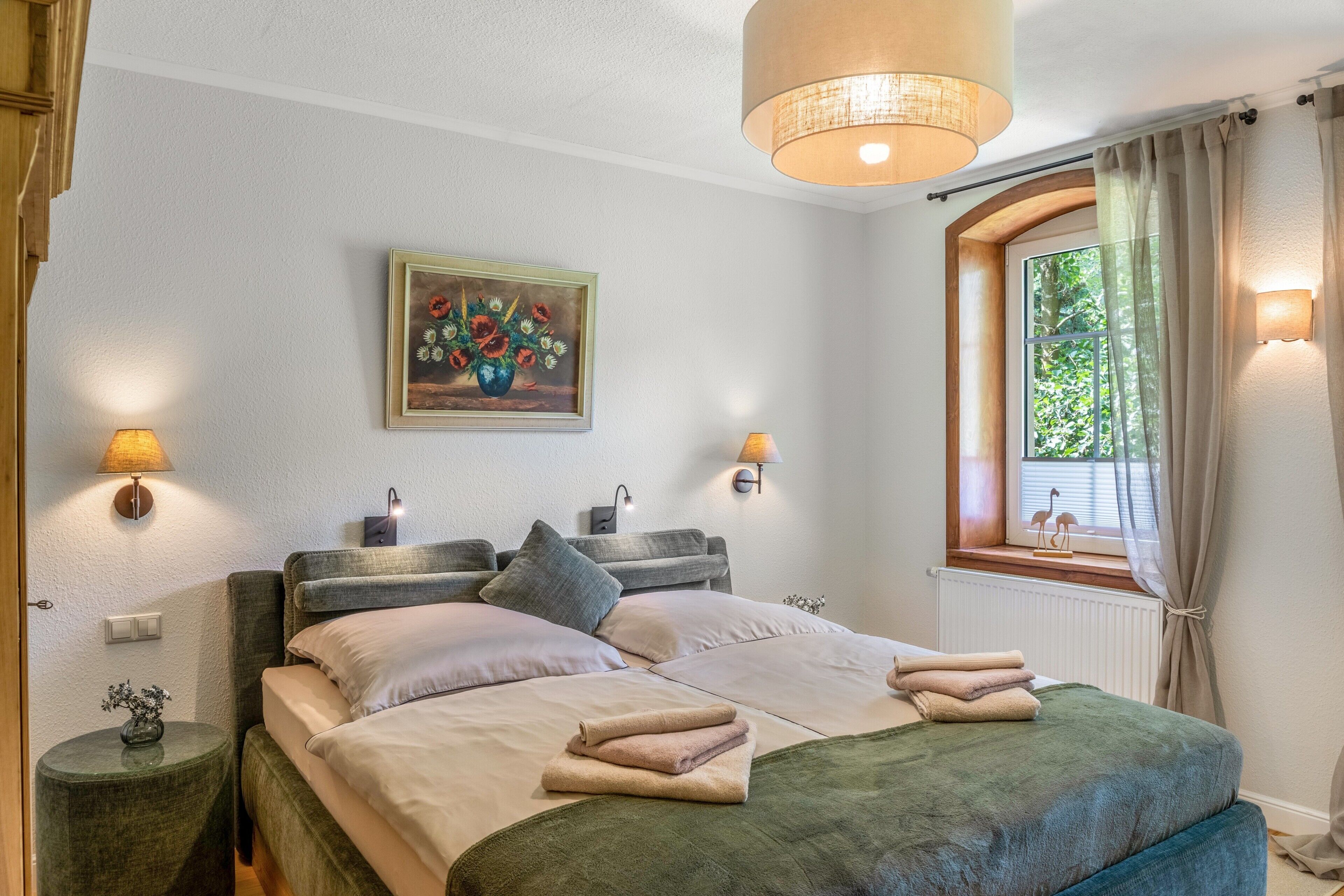Ein Schlafzimmer mit einem Bett, Nachttischlampen, einem Gemälde, einem Fenster mit Vorhängen und Blick ins Grüne.