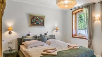 Ein Schlafzimmer mit einem Bett, Nachttischlampen, einem Gemälde, einem Fenster mit Vorhängen und Blick ins Grüne.