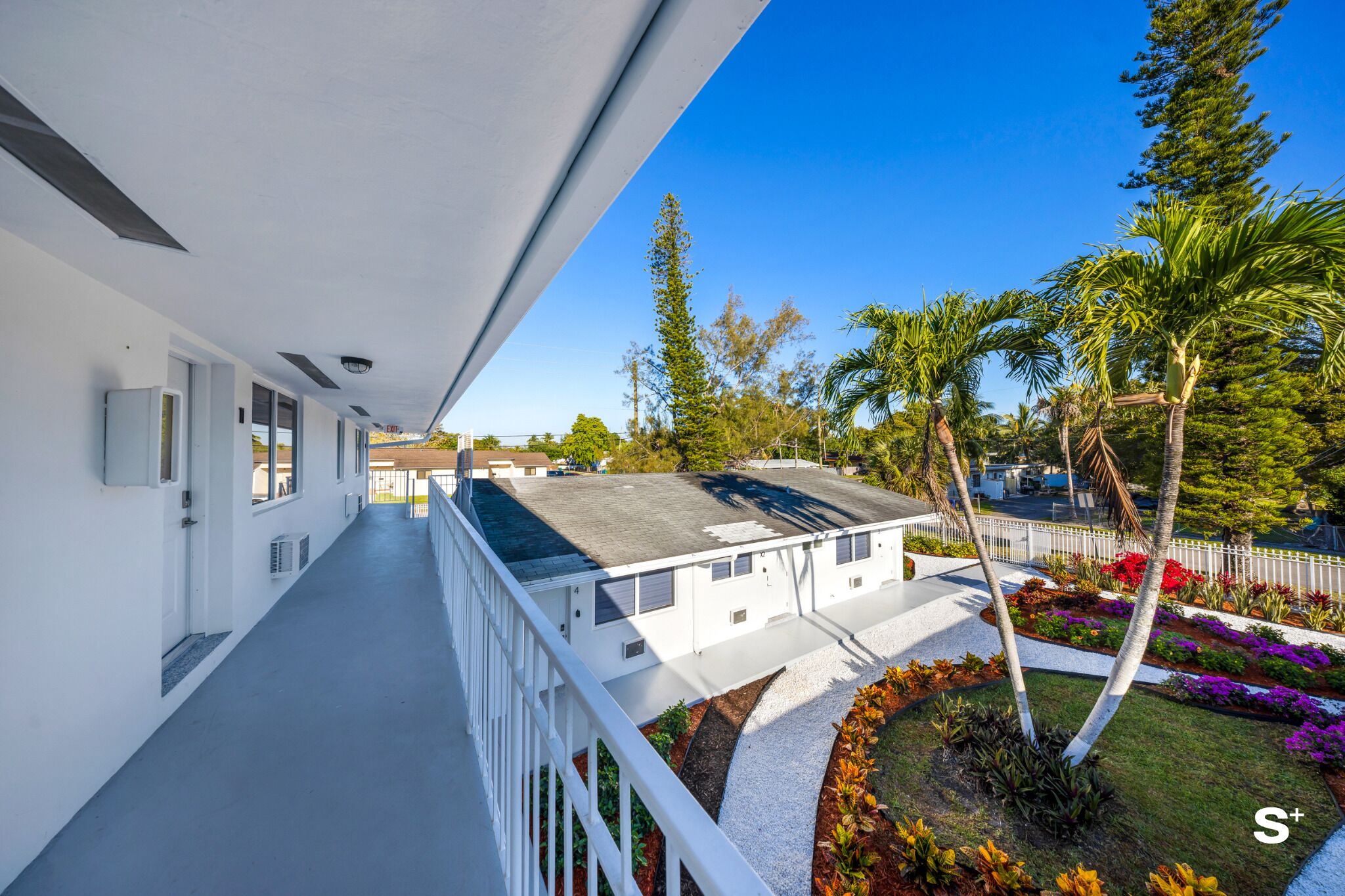 A long, narrow corridor with white walls and a balcony overlooking a landscaped area with palm trees and colorful flowers.