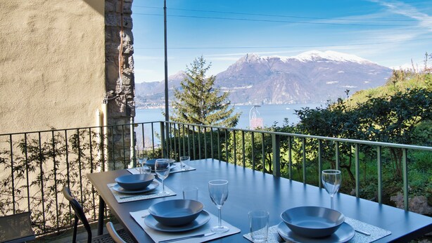 A dining table set with plates, glasses, and cutlery on a balcony with a mountain view. - Casa Rosa - hidden Garden with lake view (Fiumelatte)