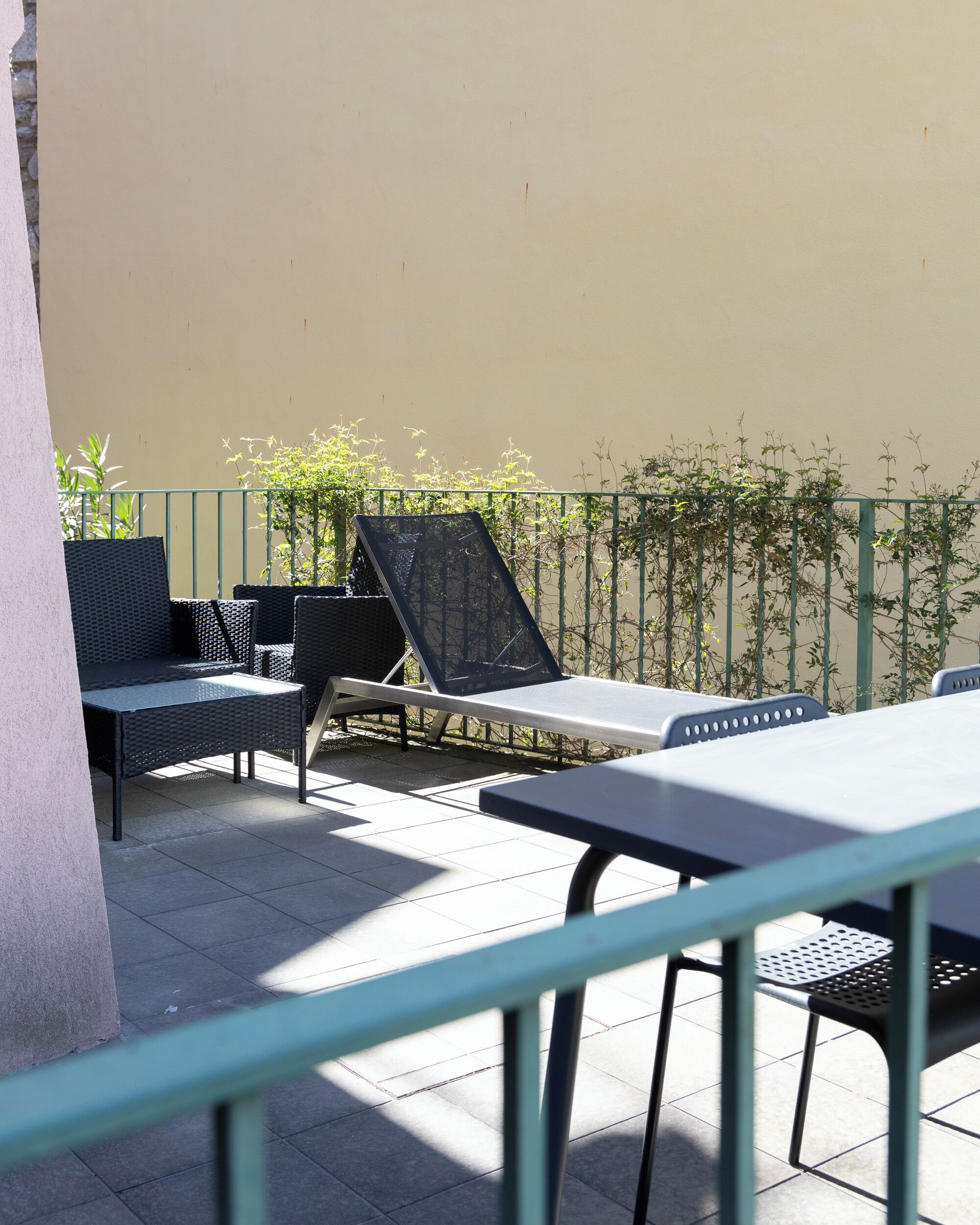 Een terras buiten met een tafel, stoelen, een ligbed en een groene metalen balustrade.