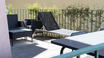 Een terras buiten met een tafel, stoelen, een ligbed en een groene metalen balustrade.