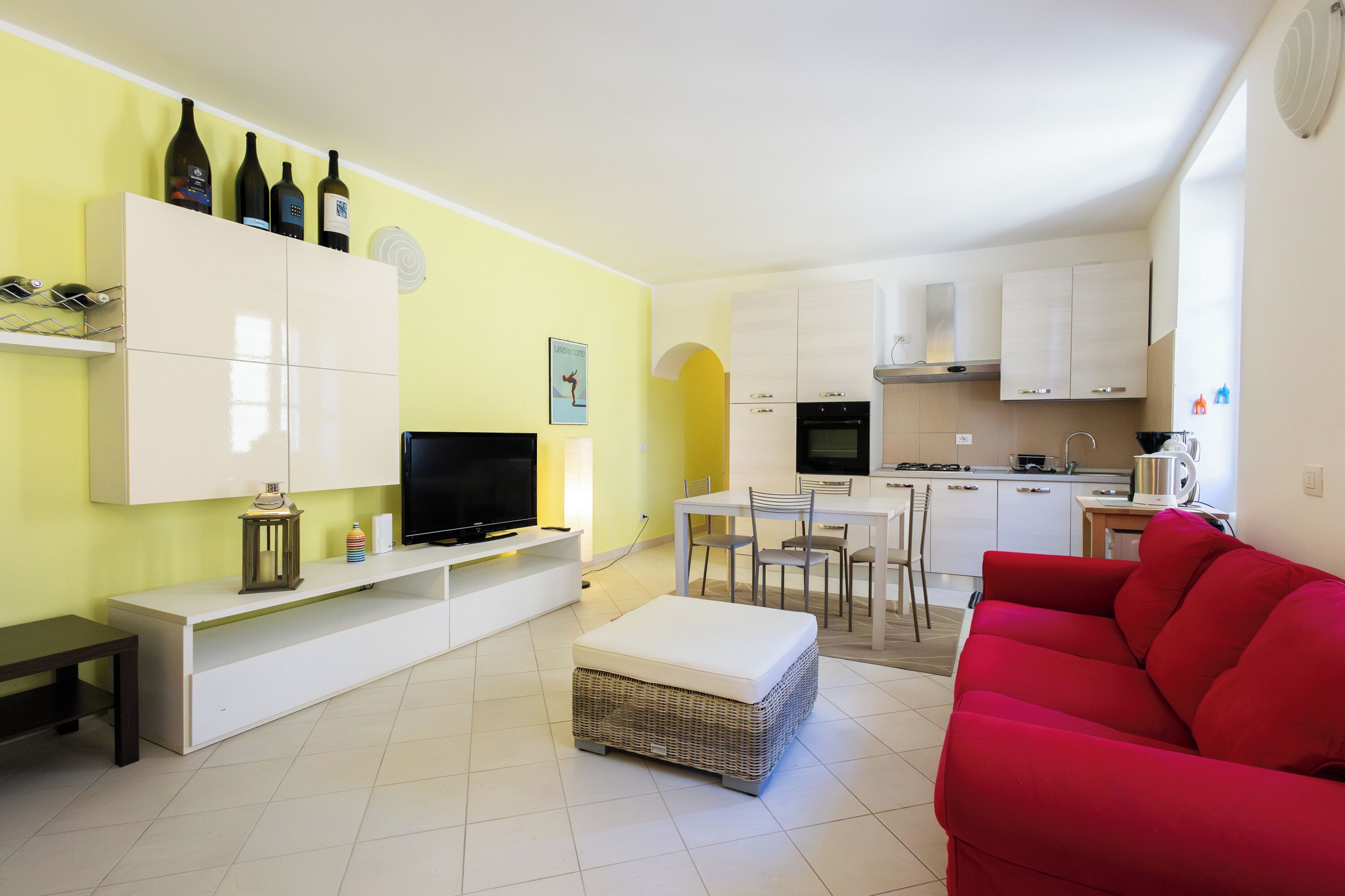 Un salon comprenant un téléviseur, un canapé et un pouf, une cuisine avec des placards et des appareils électroménagers, et un coin repas avec des chaises.