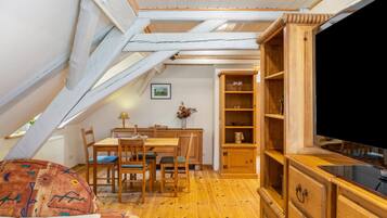 Ein gemütliches Dachzimmer mit einem Holzesstisch, Stühlen, einem Fernseher und einem Bücherregal.