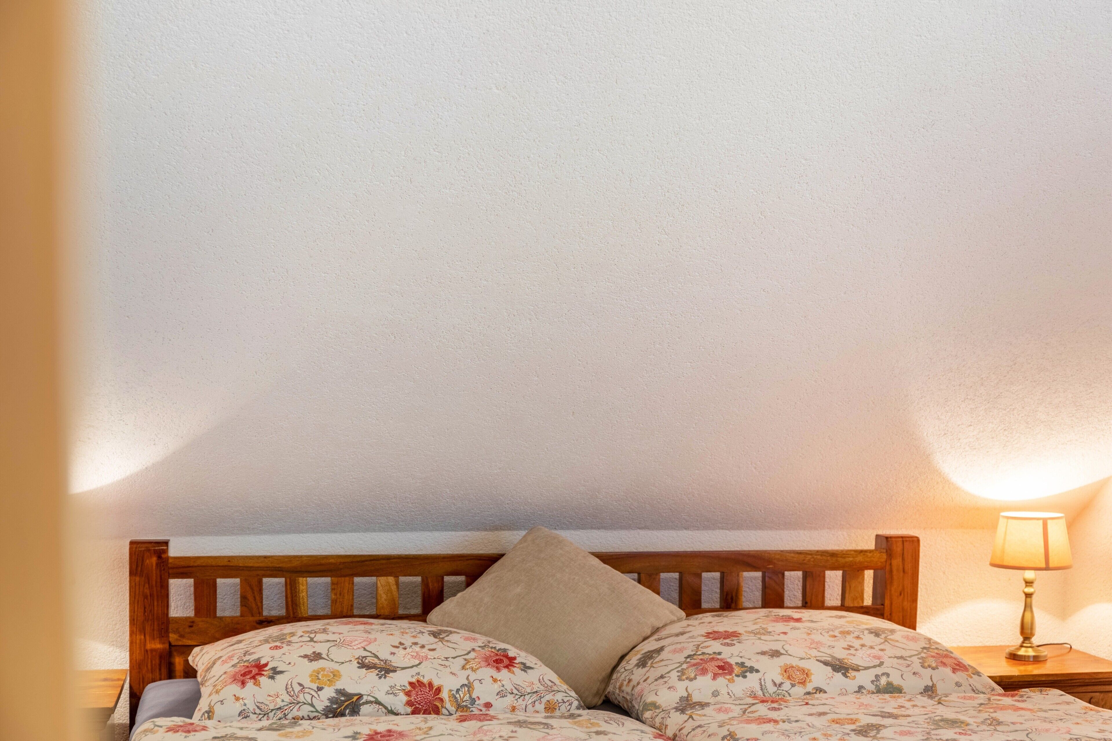 Ein Bett mit floralem Bettbezug und einem Holz-Kopfteil, eine Nachttischlampe und eine schlichte Wand.