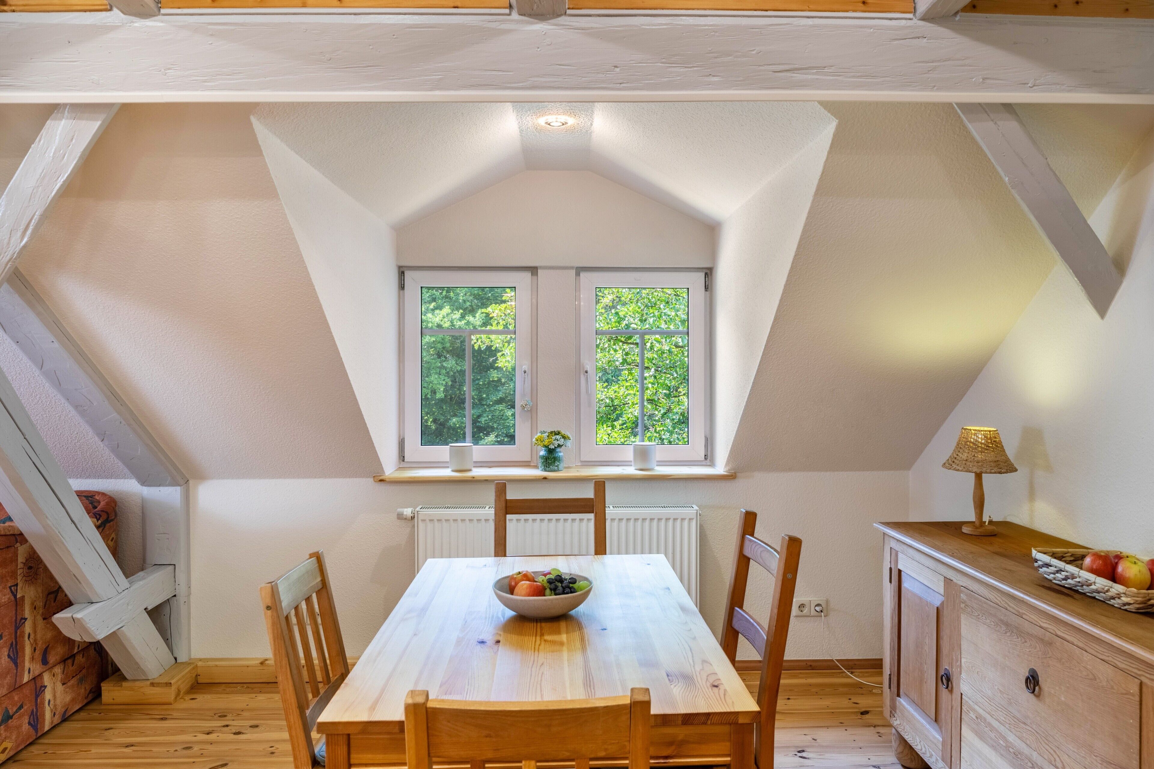 A dining area with a table, chairs, a bowl of fruit, a window with a view of trees, and a lamp on a cabinet.