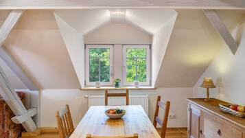 A dining area with a table, chairs, a bowl of fruit, a window with a view of trees, and a lamp on a cabinet.